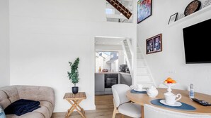 Un salon moderne avec un canapé, une petite table agrémentée d’une plante, un coin repas avec des chaises et une table préparée pour le thé, et une cuisine en arrière-plan.