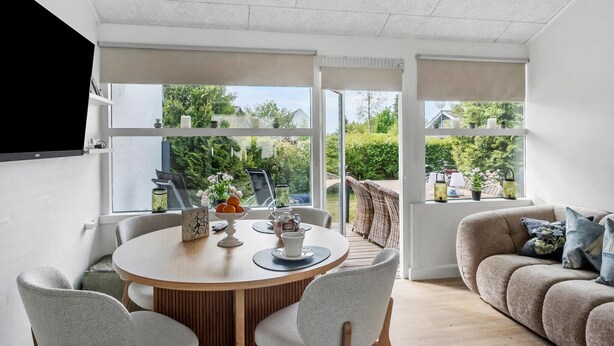 Un salon moderne avec une table à manger ronde en bois, des chaises à revêtement gris, un canapé beige et de grandes fenêtres offrant une vue sur l’extérieur.