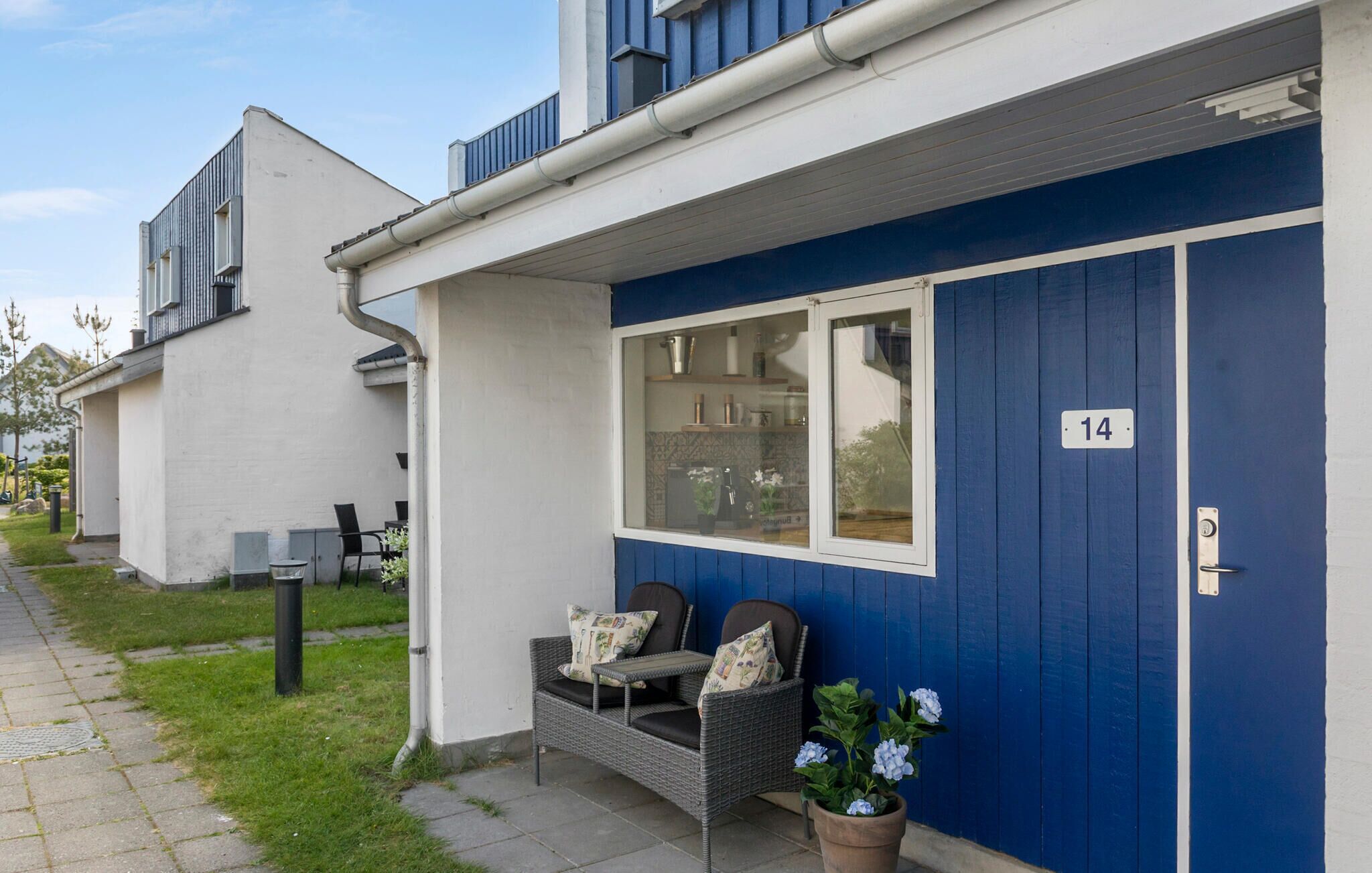 A modern building with a blue exterior, numbered 14, featuring a small outdoor seating area with wicker furniture and a potted plant.