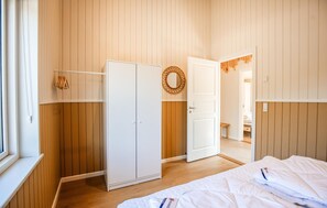 Quarto com guarda-roupa de madeira, cama com roupa de cama branca, espelho e porta para outro espaço.