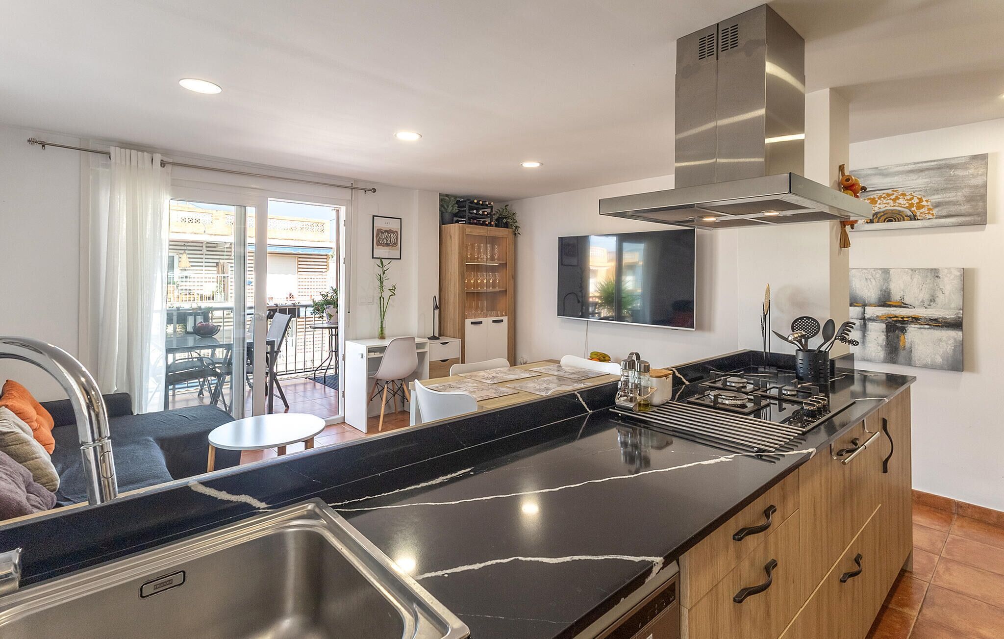 A modern kitchen with a black marble countertop, stainless steel sink, and wooden cabinets. There is a dining area with a table and chairs, and a balcony with a view.