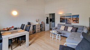 Un salon moderne avec un canapé gris, une table à manger avec des chaises et un coin cuisine visible à travers une porte ouverte.
