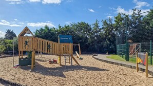 Une aire de jeux avec une balançoire en bois et un ensemble de balançoires, entourée d’une zone sablonneuse et d’un espace clos par une clôture.