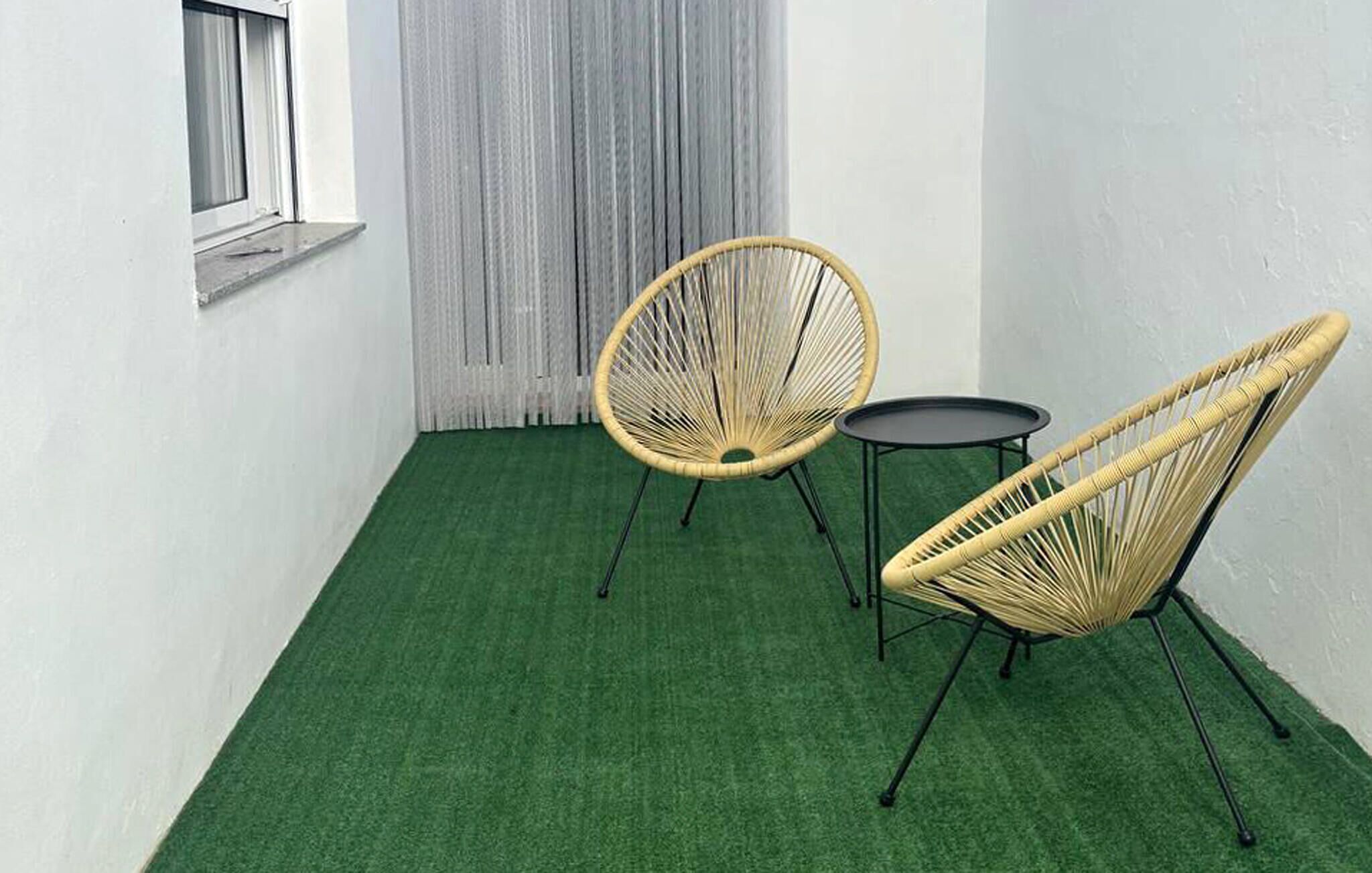 Een klein terras met twee rieten stoelen en een ronde tafel, voor een groene kunstgazon en witte muren.