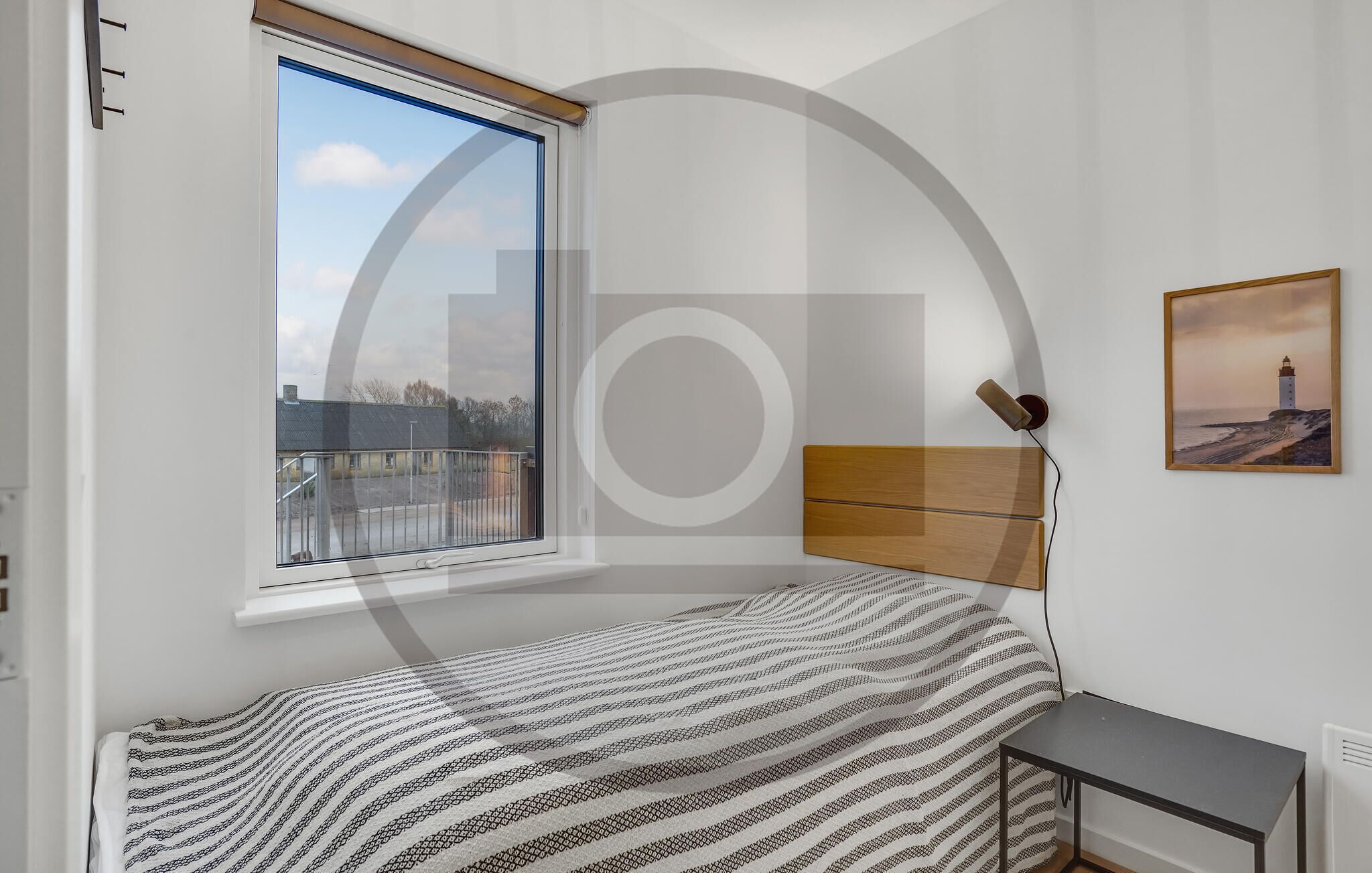 A bedroom with a bed, a small table, a lamp, a picture on the wall, and a window with a view of buildings and a fence.