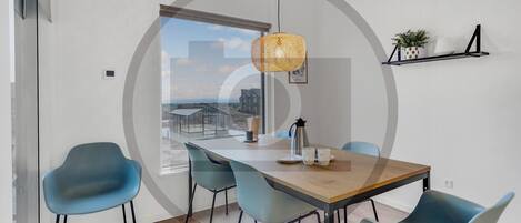A modern dining area with a wooden table, four blue chairs, a hanging light fixture, and a view of a cityscape through a large window.
