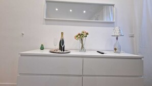Un tocador blanco con espejo, una botella de champán, una copa, un florero con flores y una lámpara.