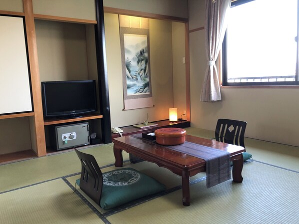 A traditional Japanese-style room with tatami flooring, a low table with cushions, a television, a painting, and a window with curtains.