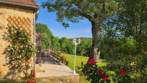 Un edificio de piedra con una pérgola de madera cubierta de rosas rosadas, un sendero de piedra que conduce a una valla blanca, y un gran árbol con rosas rojas en un jardín exuberante.