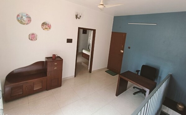 A room with a bed, a desk, a chair, and a wardrobe. There is a ceiling fan and a door leading to another room.