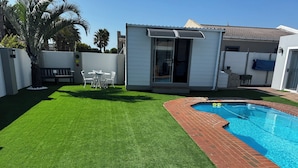 A backyard with artificial grass, a swimming pool, and a patio area with outdoor furniture. - Cosy space for work or getaway close to Blougberg beach Cape Town (Cape Town)