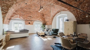 A spacious loft with exposed brick ceilings, a loft bed, a dining area with a table and chairs, and a large window providing natural light.