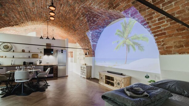 A spacious living area with a large screen displaying a tropical beach scene, a cozy seating area, and a modern kitchen with a brick ceiling.
