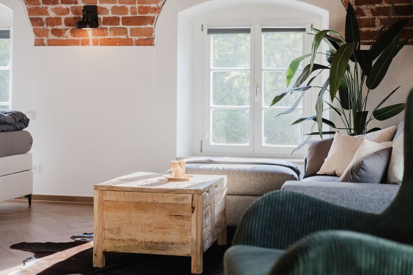 Ein gemütliches Wohnzimmer mit Backsteinwand, großem Fenster, Couchtisch aus Holz, einer Couch und einem Blumentopf.