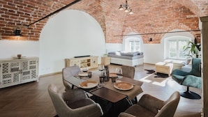 A spacious loft with exposed brick walls, a loft bed, a dining area with a table and chairs, and a cozy seating area with a sofa and armchair.