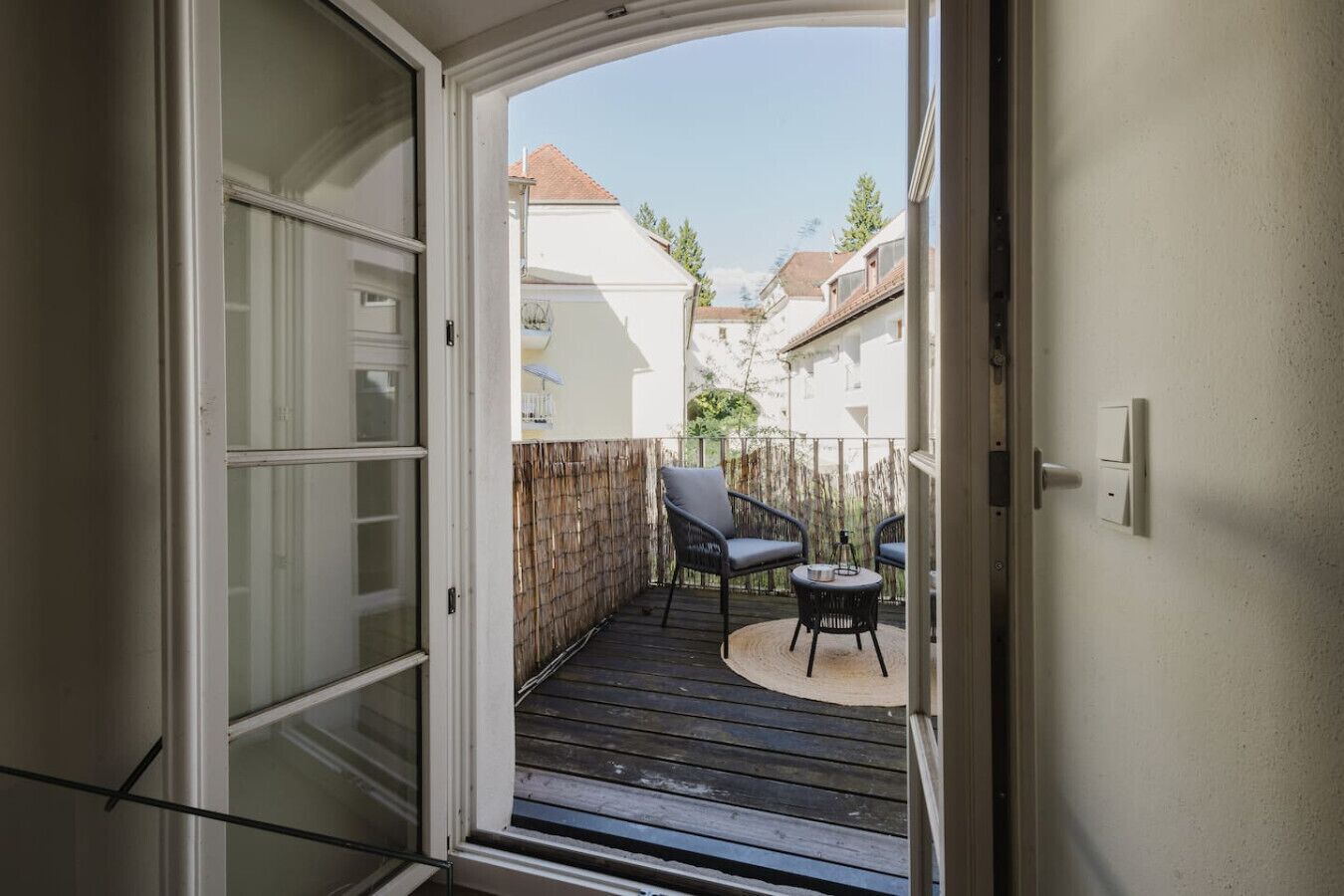 A view from a sliding glass door into a balcony with a wooden deck, two chairs, and a small table.