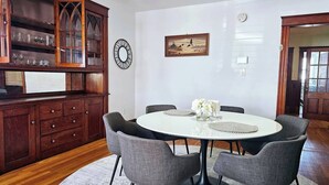 Un coin repas avec une table ronde, quatre chaises grises, un vase blanc orné de fleurs, un meuble en bois aux portes vitrées et un ventilateur de plafond.