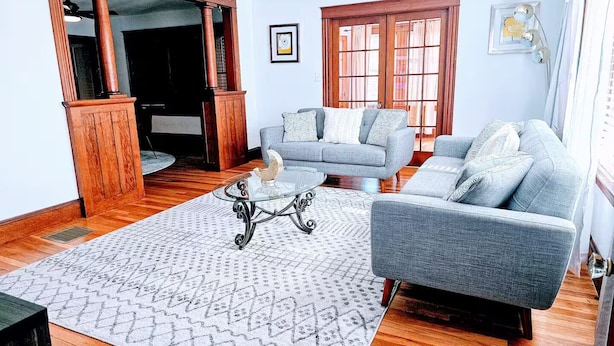 Un salon avec un canapé gris, une table basse en verre et un tapis à motifs. On y trouve également un meuble en bois et un tableau encadré accroché au mur.