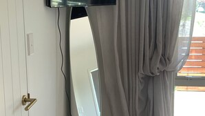 Habitació amb televisió penjada, un mirall gran, una cadira de fusta i una finestra amb cortines.
