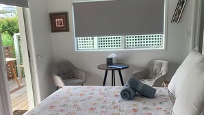 Un dormitori amb un llit gran, una taula petita, dues cadires, una finestra amb persianes i una porta que dona a una zona exterior.