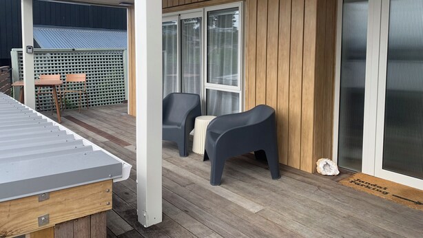 Een overdekt buitengebied met een houten terras, twee stoelen, een tafel en een schuifdeur van glas.