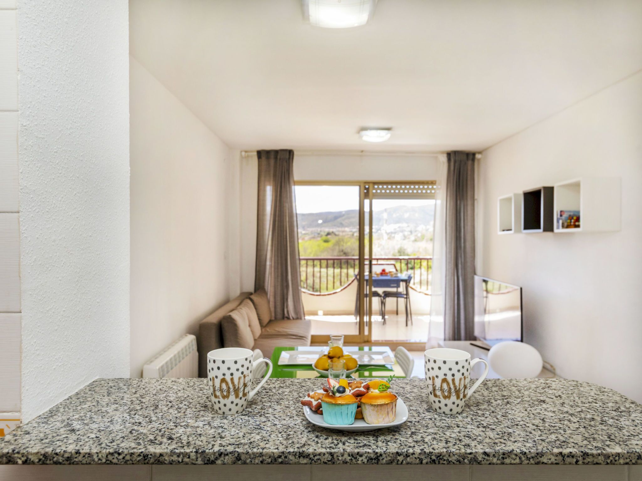A modern kitchen with granite countertops, two coffee mugs with "LOVE" written on them, and a small table with a plate of pastries.