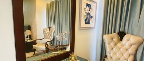 A room with a large mirror, a tufted armchair, a wooden desk, and a framed artwork on the wall.