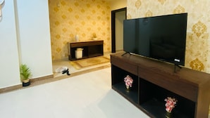 A living room with a flat-screen TV on a wooden stand, patterned wallpaper, and a tiled floor.