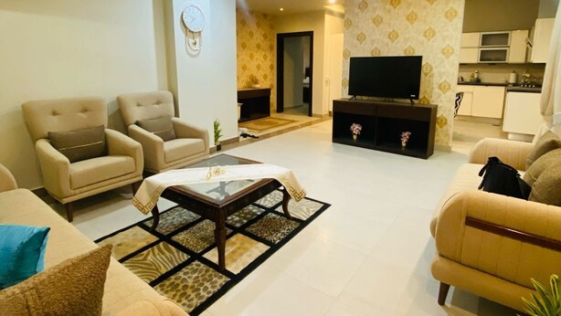 A spacious living room with a sofa set, a coffee table, and a television. There is a kitchen area in the background.