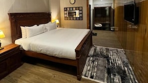 A hotel room with a wooden bed, a nightstand, a wall clock, a television, and a patterned rug. - Infinity Villa and Resort Kumrat Valley (Kumrat Valley)