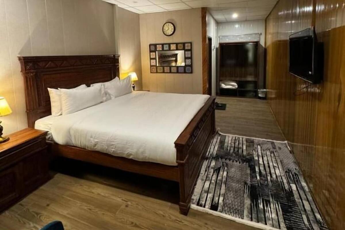 A hotel room with a wooden bed, a nightstand, a wall clock, a television, and a patterned rug.