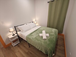 A neatly made bed with white linens and a green blanket, a bedside table with a lamp, and a green curtain.