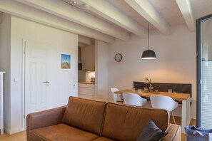Una sala de estar con un sofá marrón, un comedor con mesa de madera y sillas blancas, una cocina al fondo y un reloj en la pared.