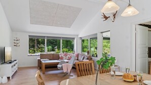 Un salon moderne avec une table à manger en bois, un ensemble de canapés et une grande fenêtre donnant sur un paysage verdoyant.