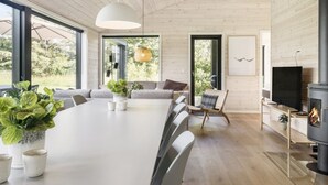 A modern living room with a large white table, comfortable seating, a fireplace, and large windows offering a view of greenery. - "Faidra" - 1.1km from the sea by Interhome (Hirtshals)