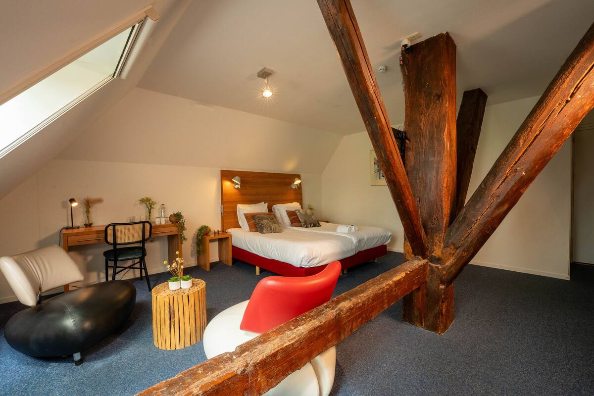 Ein Schlafzimmer mit schrägem Dach, einem Bett, einem Schreibtisch, einem Stuhl und einem roten Hocker.