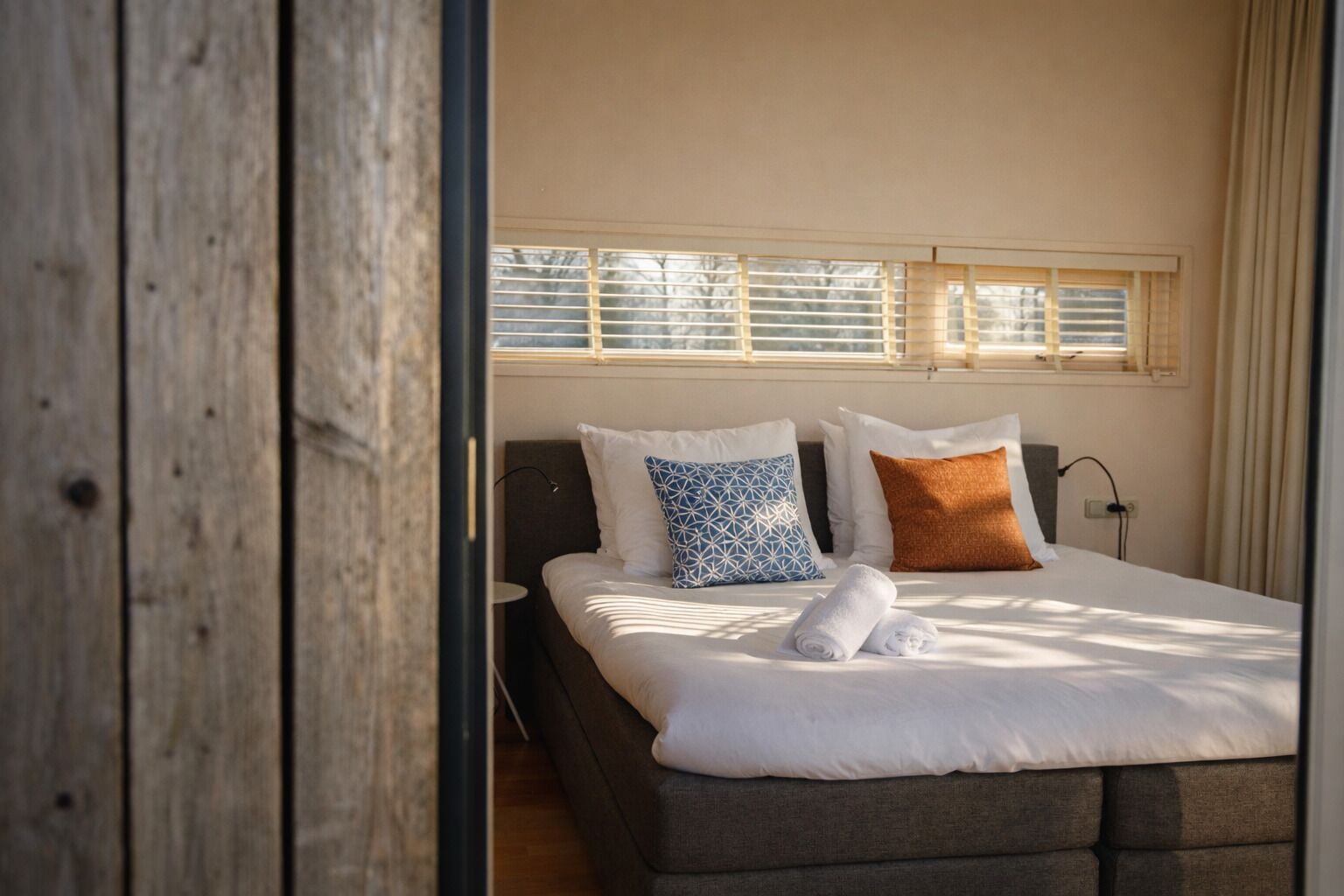 A bedroom with a bed, pillows, a bedside table, and a window with blinds.