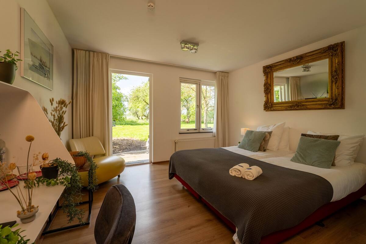 Ein Schlafzimmer mit Bett, Schreibtisch, Stuhl, großem Spiegel und Blick ins Freie.