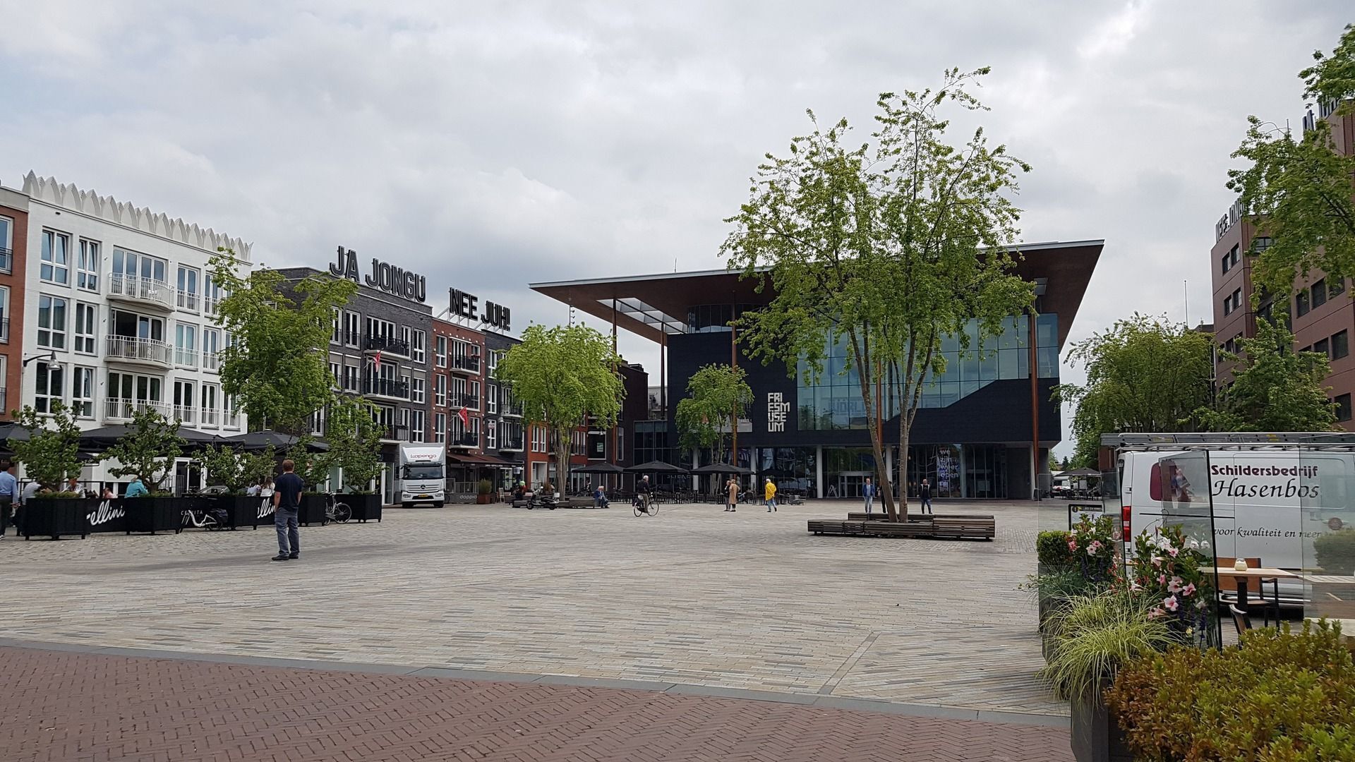 A public square with a modern building, trees, and a white van with text.