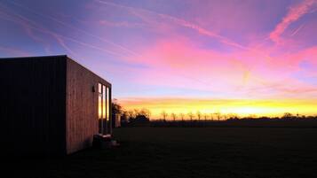 A wooden building with a large window, a grassy field, and a sunset sky with pink and orange hues.