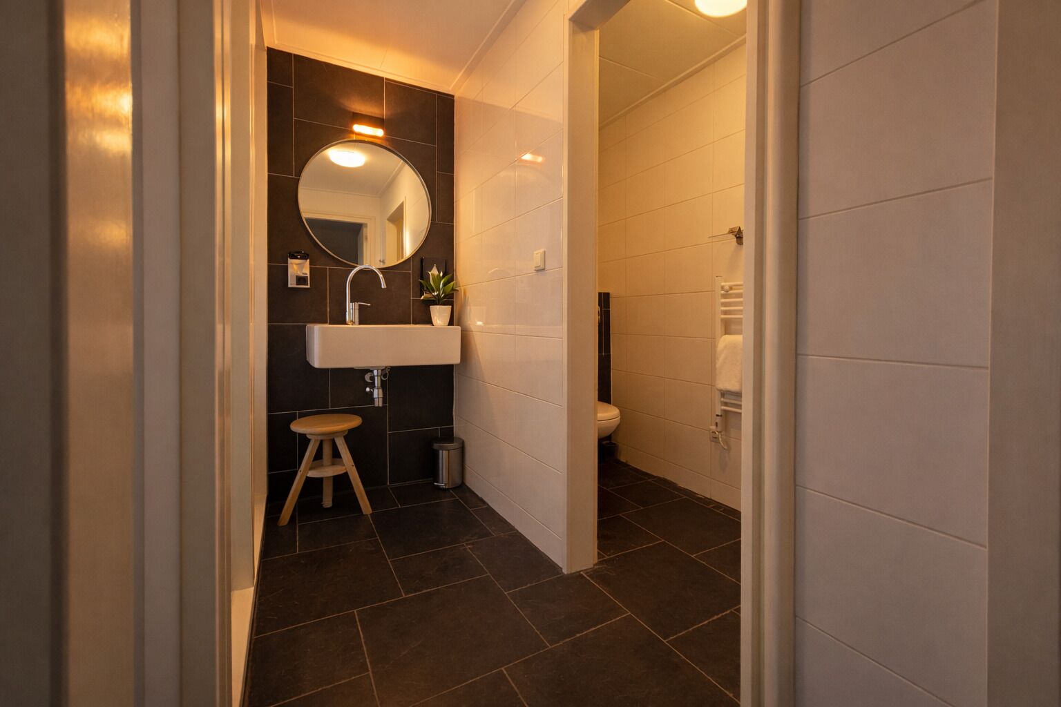 A modern bathroom with a dark tiled floor, a white sink with a round mirror, a small stool, and a white toilet.
