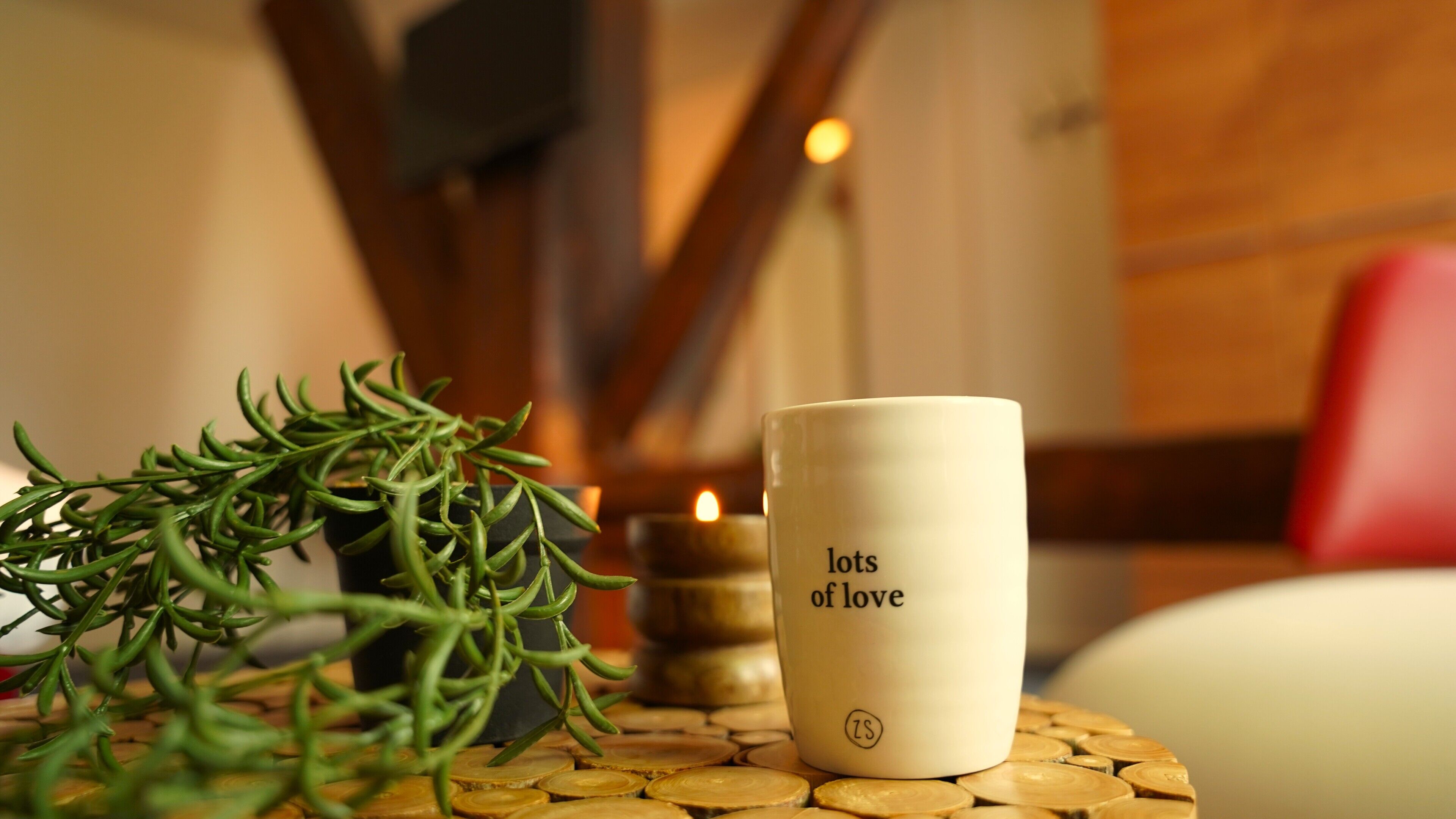 A white mug with "lots of love" text, a green plant, and a lit candle on a wooden table.