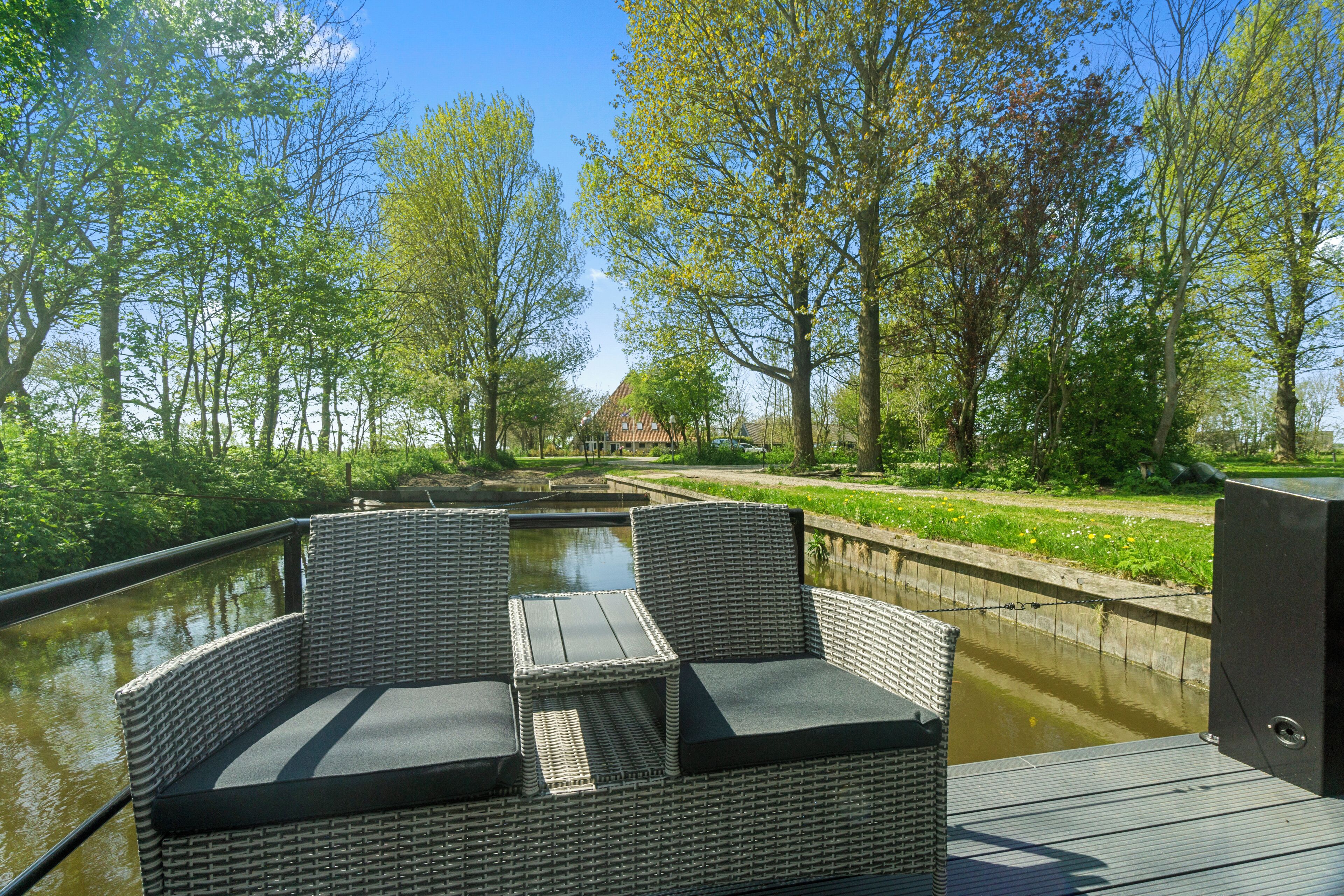 A wicker lounge chair with cushions by a canal, surrounded by trees and a house in the background.