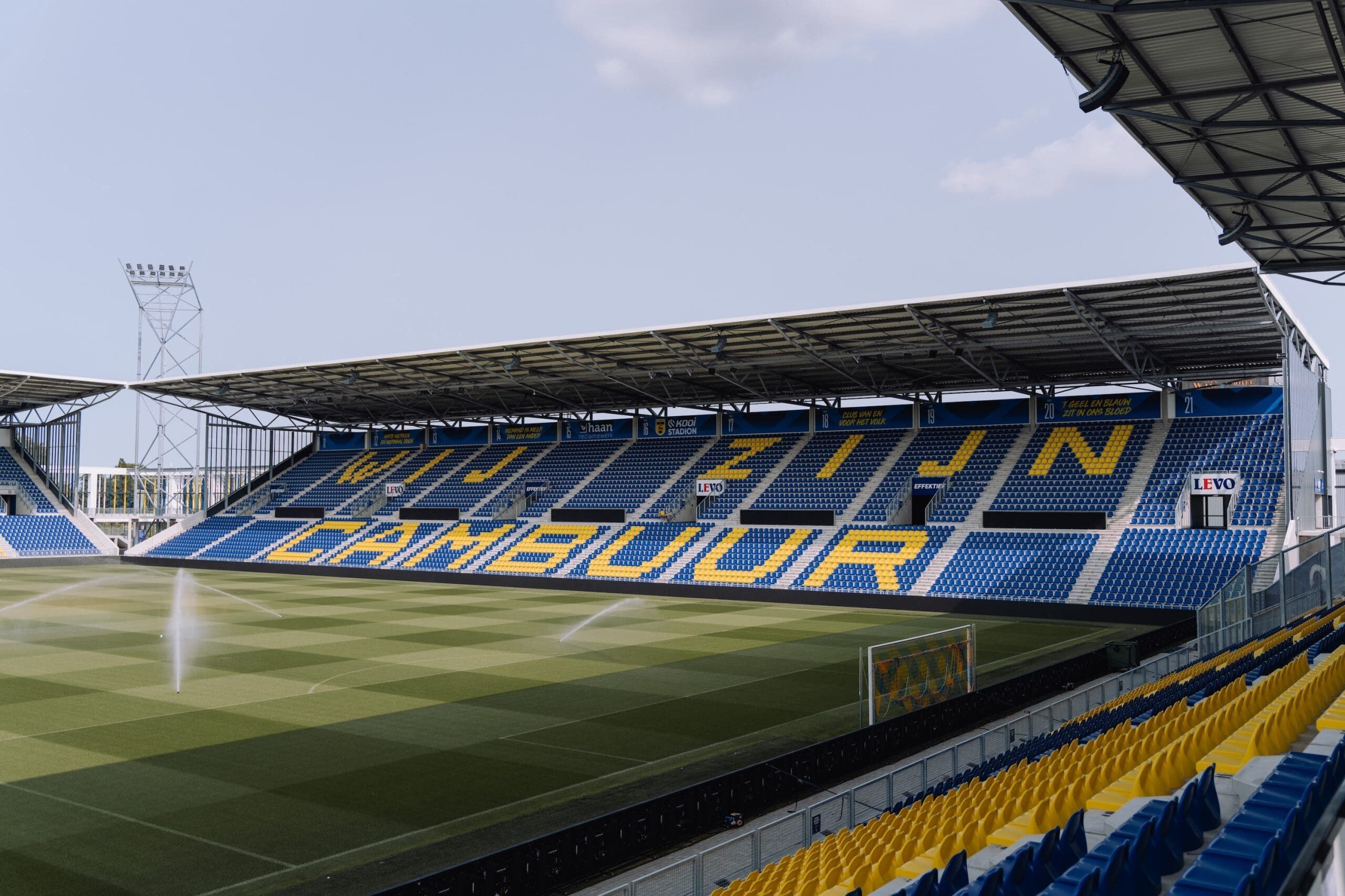 A stadium with blue and yellow seating, a roof structure, and a visible scoreboard.