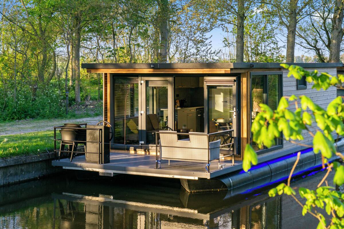 Ein schwimmendes Hausboot mit einer Terrasse, einer Sitzgelegenheit und einer durch die Glastüren einsehbaren Küche.