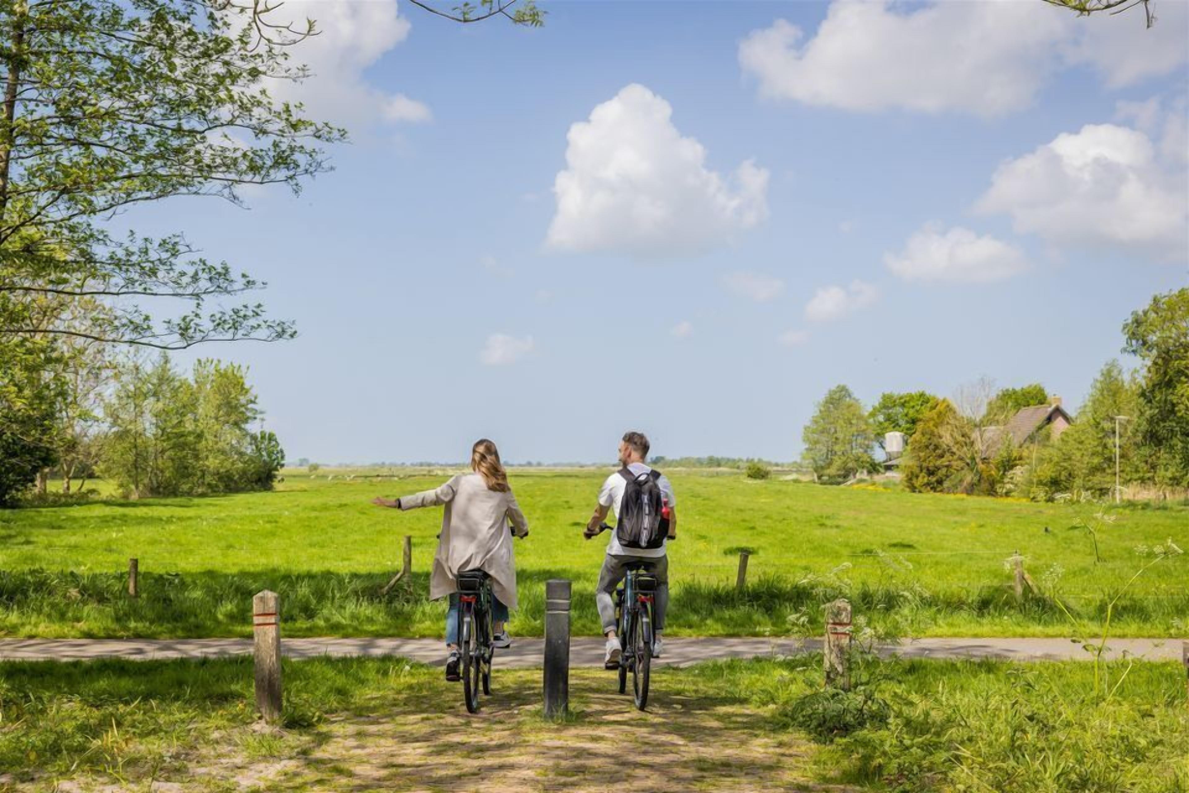 Två personer cyklar på en stig i ett lantligt område med gröna fält och en klar himmel.