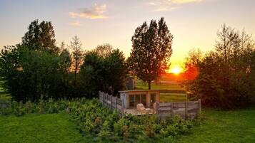 A wooden cabin with a deck and a chair, surrounded by trees and a grassy field, with the sun setting in the background.