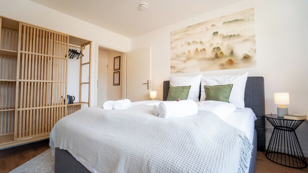 A neatly arranged bedroom with a large bed, a wooden wardrobe, and a wall-mounted artwork.