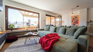 A modern living room with a large window, a green sofa, a red throw blanket, and a patterned rug.
