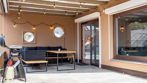 An outdoor patio with a wooden table and benches, string lights, and a grill.